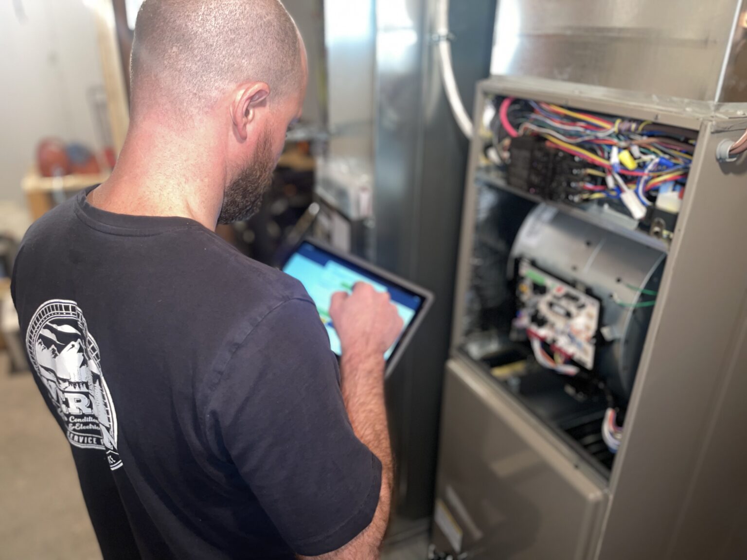 HVAC technician tapping a tablet in front of an open gas furnace with the control board and wiring exposed